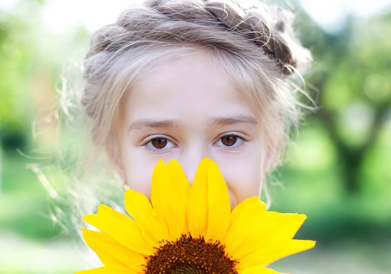 A young girl with a braided hairstyle peeks over a bright yellow sunflower in a sunny garden.