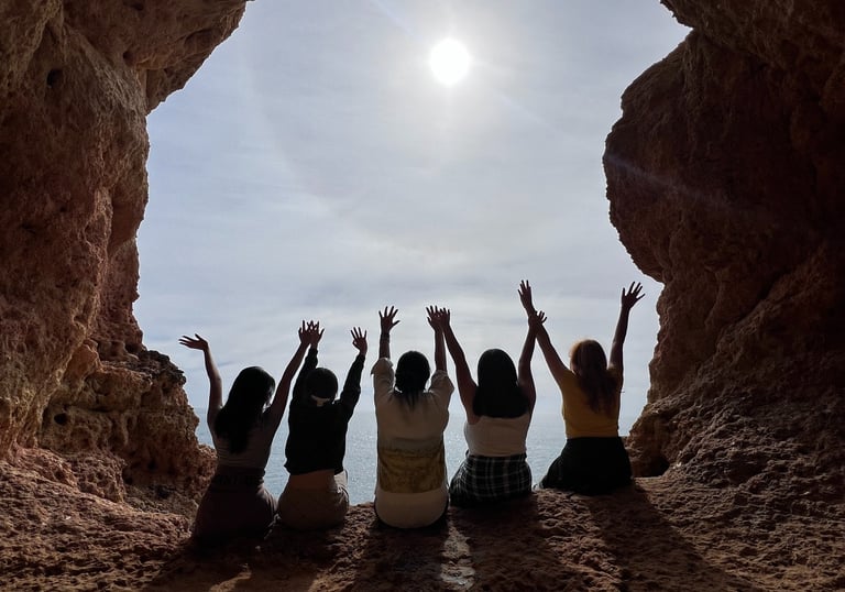 Five friends raising their arms inside a sea cave overlooking the bright sun and ocean in Algarve.