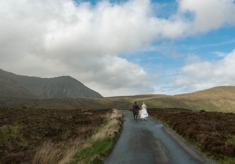 A bride and groom in a kilt walk down a road in the scenic Isle of Skye mountains.
