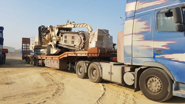 تريلا لوبد زرقاء تحمل حفارًا كبيرًا بلون البيج/البني الفاتح، وهي جاهزة لخدمات نقل معدات ثقيلة.