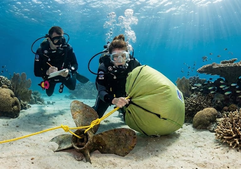 Scuba divers using a yellow lift bag to recover a sunken boat propeller from a coral reef seabed.