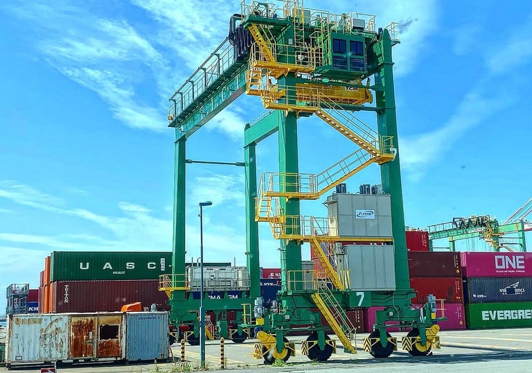 A large green gantry crane standing over shipping containers at a busy commercial port terminal.