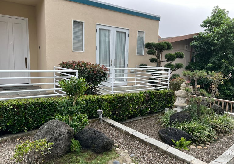 Modern Japanese zen garden with a stone lantern, manicured shrubs, and lava rocks in front of a tan house.