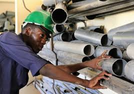 A plumber in a green hard hat organizes metal pipes on a storage rack in a warehouse.
