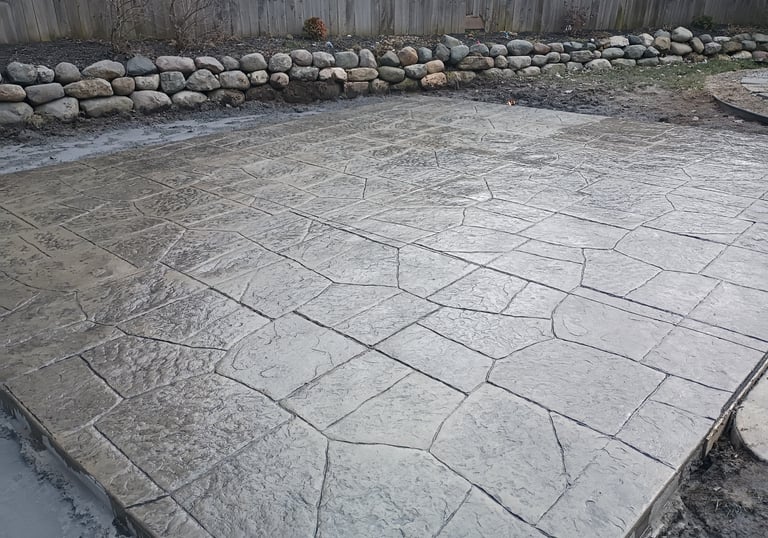 Freshly poured gray stamped concrete patio with flagstone pattern and rock wall landscape border.