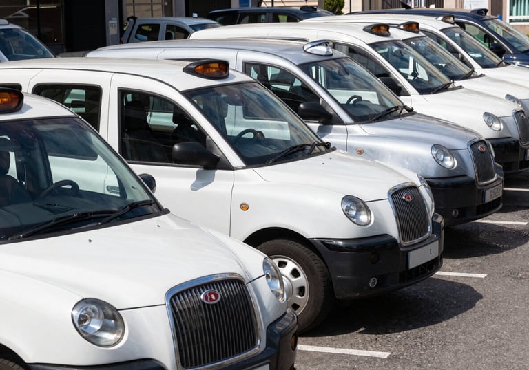 A row of clean, professional white and silver taxis parked neatly at a European / British transit hub. Clear, bright daylight, emphasizing organization and readiness.