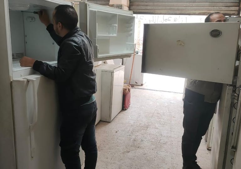 Professional technician in a black jacket repairing a white refrigerator in an appliance service shop.