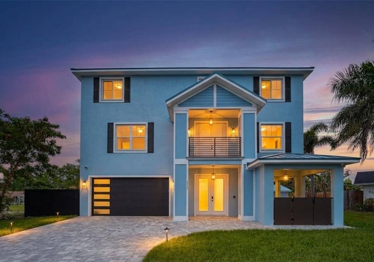A modern light blue two-story home with a paved driveway and warm exterior lighting at sunset.
