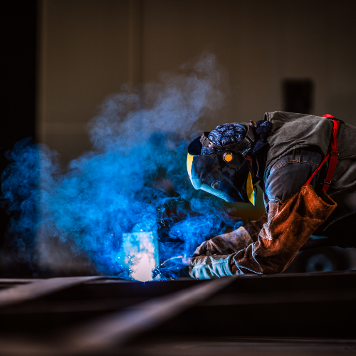 Professional welder in protective gear and mask performing metal fabrication with bright blue sparks and smoke.