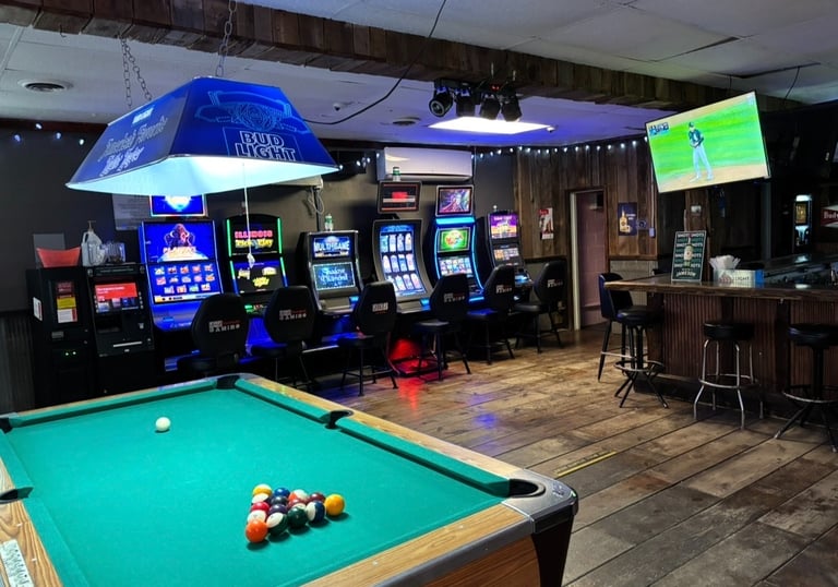 Rustic sports bar interior with a green pool table, neon slot machines, and a wooden bar area.