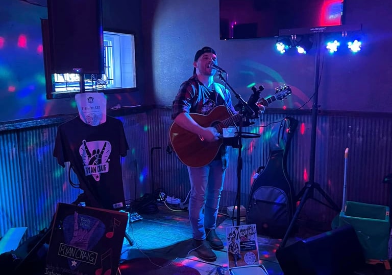 Musician Ryan Craig playing acoustic guitar and singing on a small stage with blue and purple disco lights.