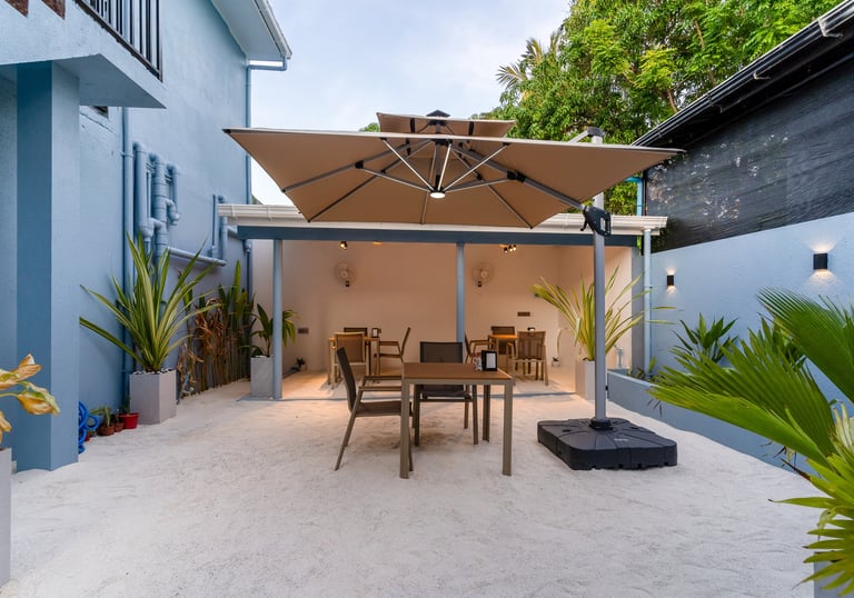 Modern outdoor cafe seating area with a large tan umbrella, dining tables, and tropical plants.