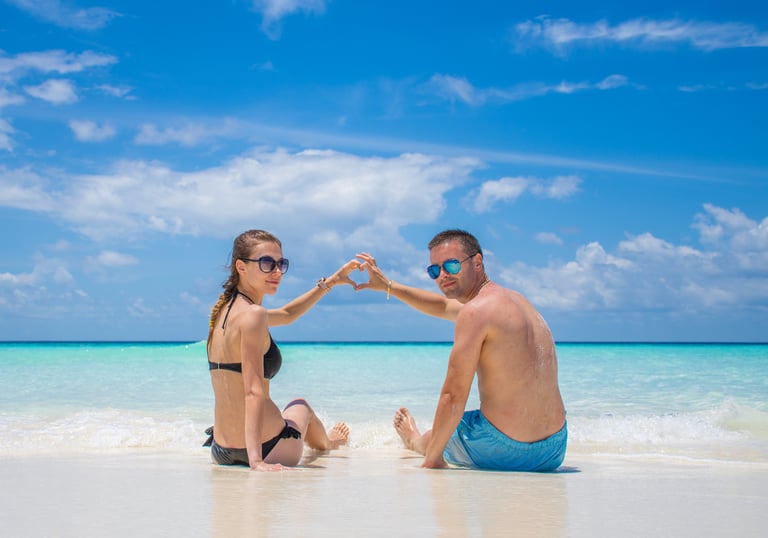 A couple making a heart shape with their hands on a tropical beach with turquoise water.