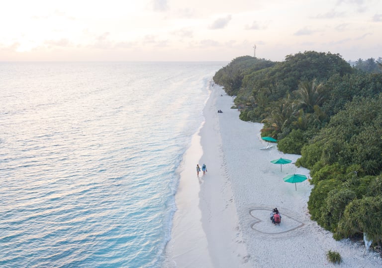 Aerial view of a couple walking on a white sand beach at sunset in the Maldives, with lush green trees and turquoise water.