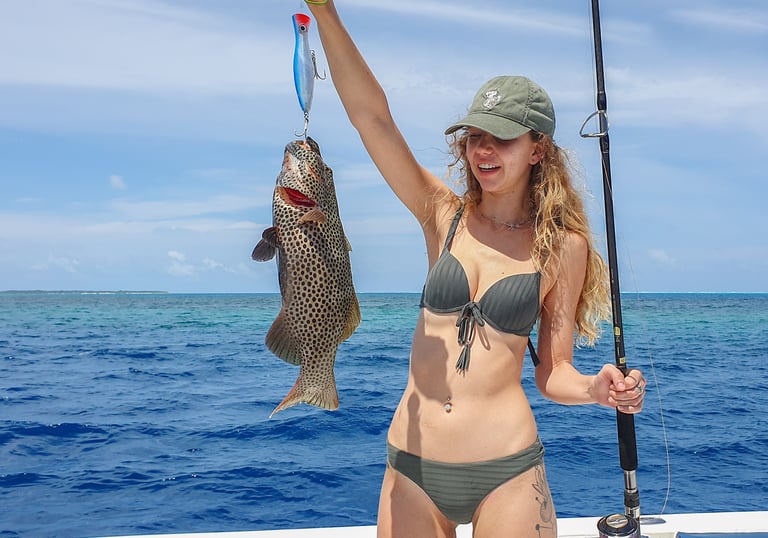 A smiling woman on a boat holds up a large spotted grouper fish caught on a blue lure while deep sea fishing.