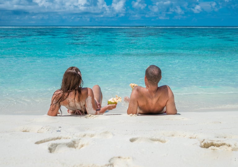 A couple relaxing on a white sand beach in the Maldives with coconut drinks by turquoise water.