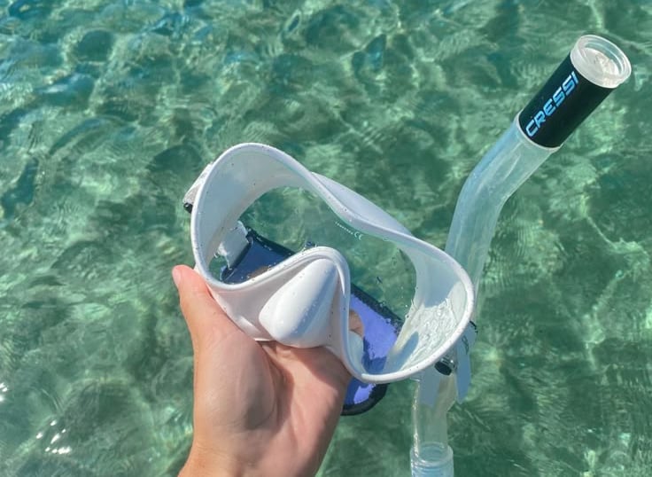 A hand holding a white Cressi snorkeling mask and snorkel over clear turquoise ocean water.