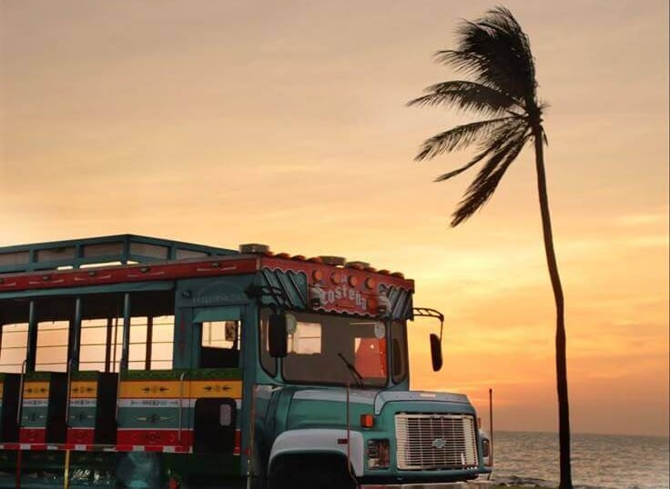 A colorful open-air Chiva bus drives along a coastal road at sunset with a palm tree.