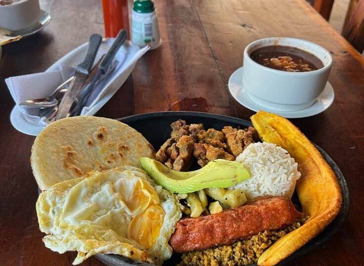 Traditional Colombian Bandeja Paisa with fried egg, avocado, plantain, rice, and beans on a rustic wooden table.