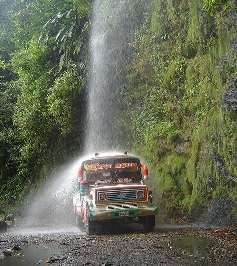 A colorful vintage Transur bus driving under a natural waterfall on a narrow jungle road.