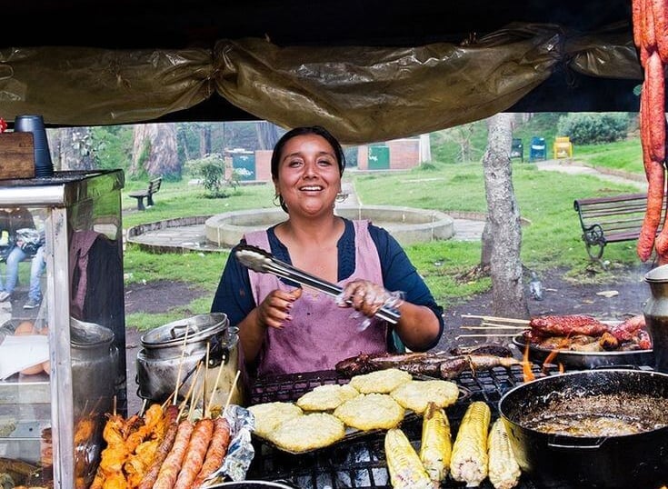 A smiling street food vendor grilling corn on the cob, sausages, and arepas at an outdoor market.