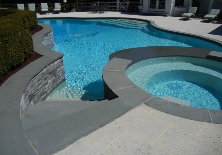 Curved pool with circular spa, light blue water and grey concrete coping with pale interior tiles.