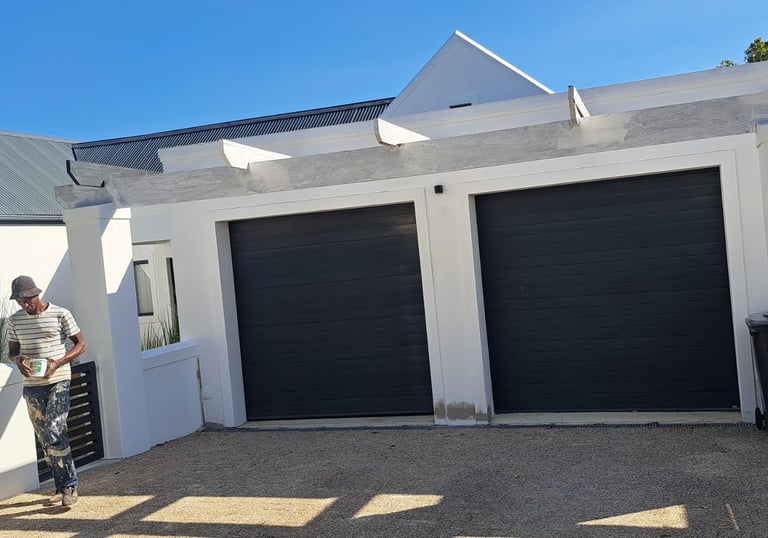A professional painter applying a fresh coat of white paint to a modern house with two black garage doors.