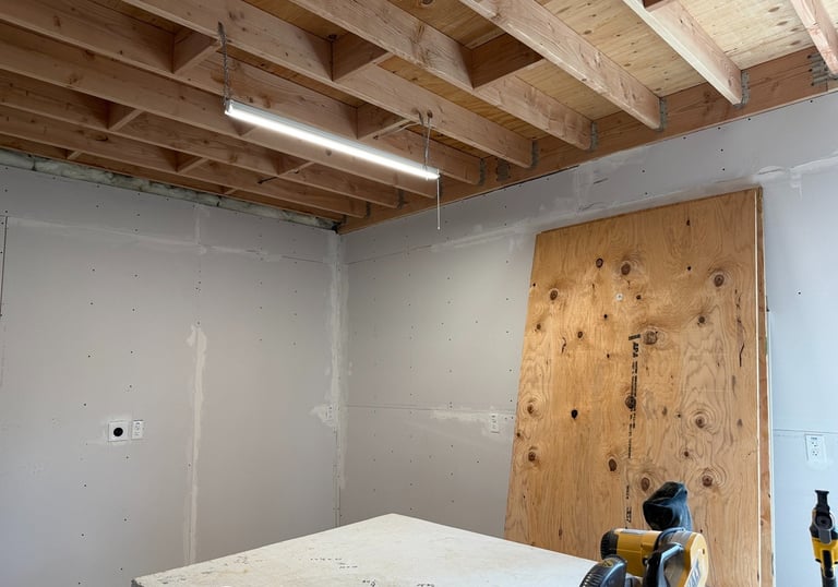 Basement renovation progress with exposed ceiling joists, drywall installation, and shop lighting.