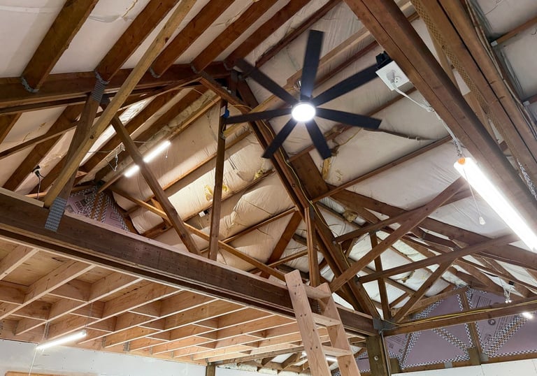Exposed wooden roof trusses in a garage attic with a black ceiling fan and bright LED lights.