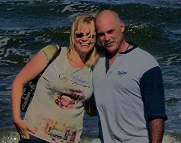 A smiling couple posing together on a beach with ocean waves in the background. Portsmouth, VA