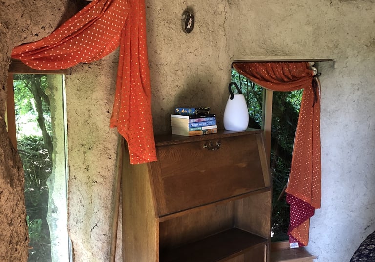 interior of a cob round-house showing a bureau, orange curtains and a pile of books