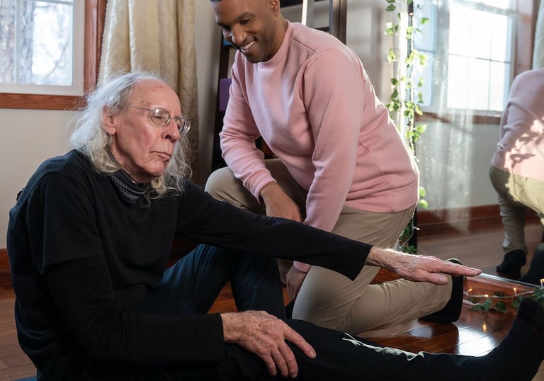 A caregiver assists an elderly man with leg stretches on a yoga mat during a home session.