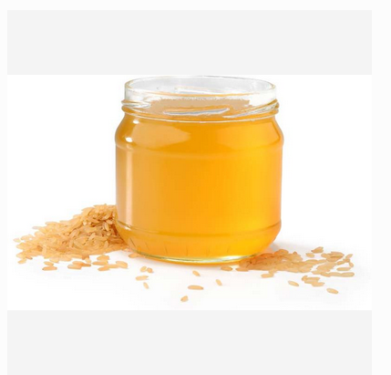 A glass jar of yellow rice bran oil surrounded by scattered grains of brown rice on a white background.