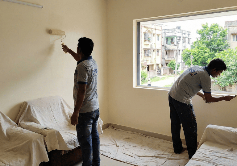 Professional painters applying fresh beige paint to living room walls with a roller and brush.