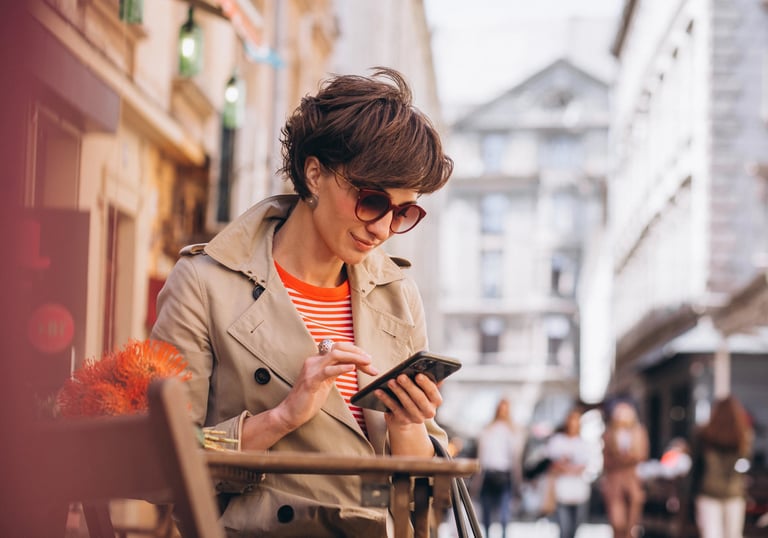 Stilinga moteris su smėlio spalvos lietpalčiu ir akiniais naudojasi telefonu lauko kavinėje.