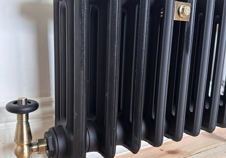 Vintage-style black cast iron radiator with brass thermostatic valves on a light wood floor.