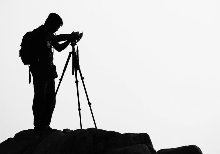 Silhouette of a travel photographer with a backpack using a tripod and camera on a rocky peak.