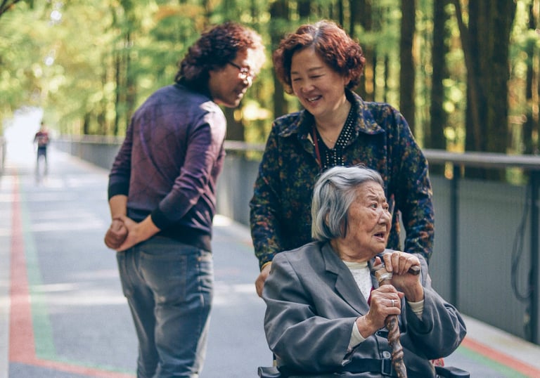 A smiling woman pushes an older woman with grey hair in a wheelchair while a man walks past