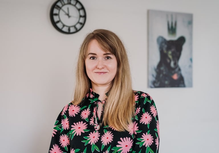 Psichoterapeutė Evelina A. stovinti šviesiame kambaryje; fone matosi sieninis laikrodis, paveikslas