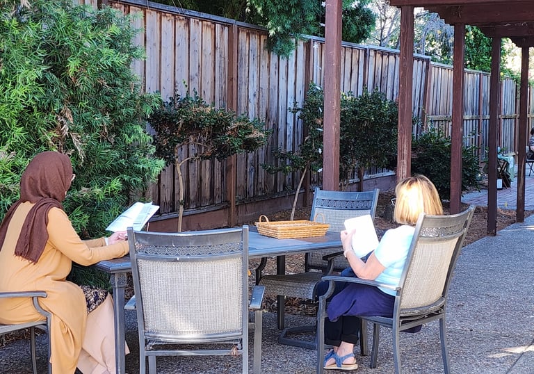 Two women at a table outside reading. One with a hijab, one with light hair.