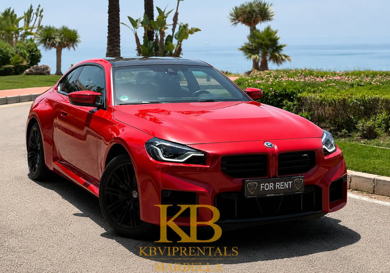 Red BMW M2 rental car parked on a sunny Marbella coastal road with palm trees and ocean view.