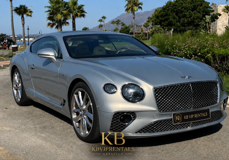 Silver Bentley Continental GT luxury car rental parked in Marbella with palm trees and mountains.