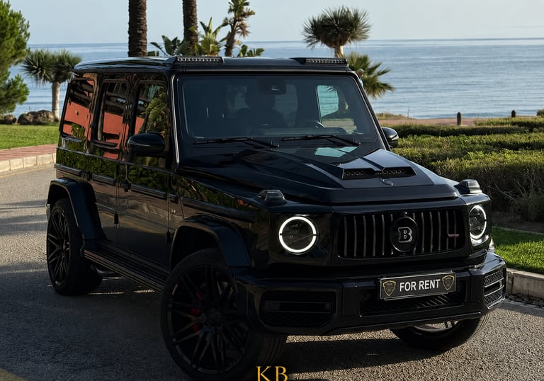Luxury black Brabus G-Wagon rental parked by the sea in Marbella with palm trees.