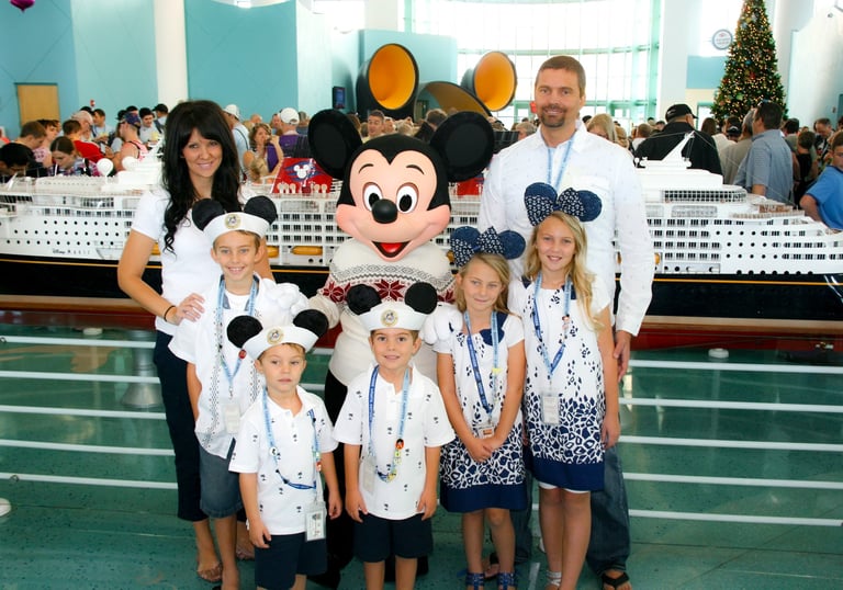 A family poses with Mickey Mouse in front of a Disney Cruise Line ship model at a port terminal.