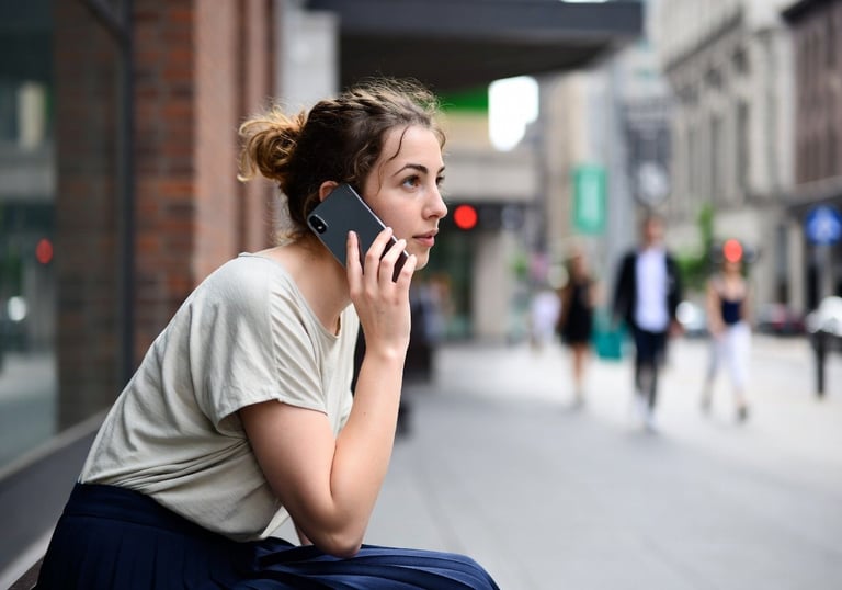 Young woman talking on a smartphone while sitting on a city street sidewalk.