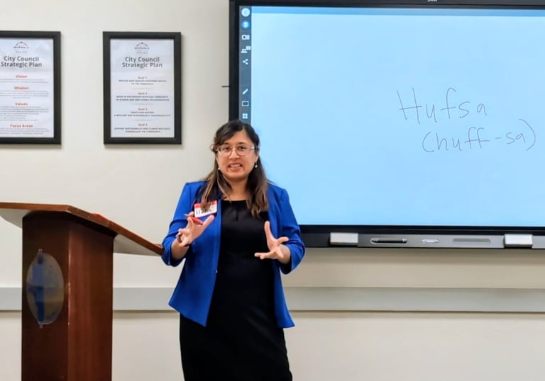 A woman in professional clothing with a blue blazer is speaking in front of a whiteboard, two awards, and a podium.