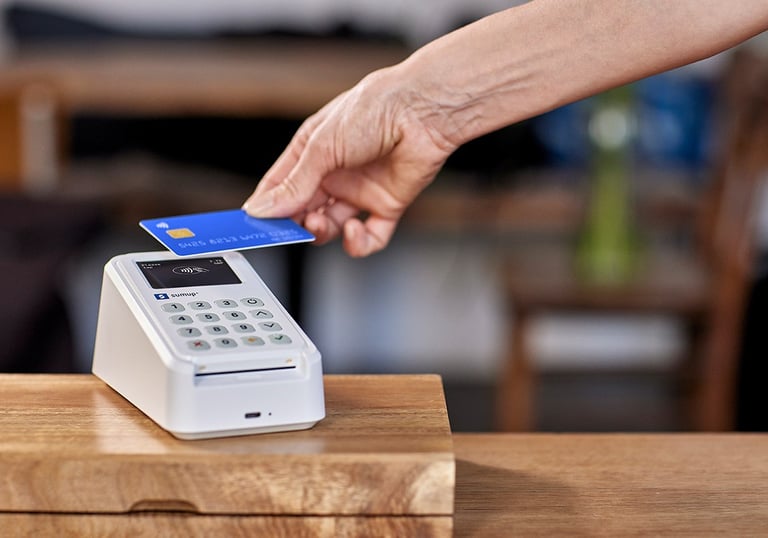 Customer making a contactless credit card payment on a white SumUp card reader at a retail counter.