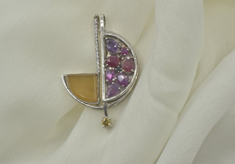A silver geometric pendant featuring amethyst, ruby, and citrine gemstones on a white fabric background.