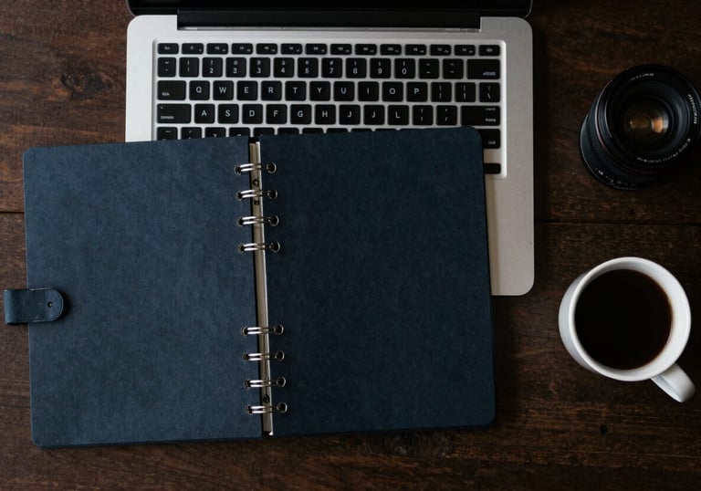 A top-down view of a minimalist North American / US workspace with a laptop, a dark navy planner, and a cup of coffee.