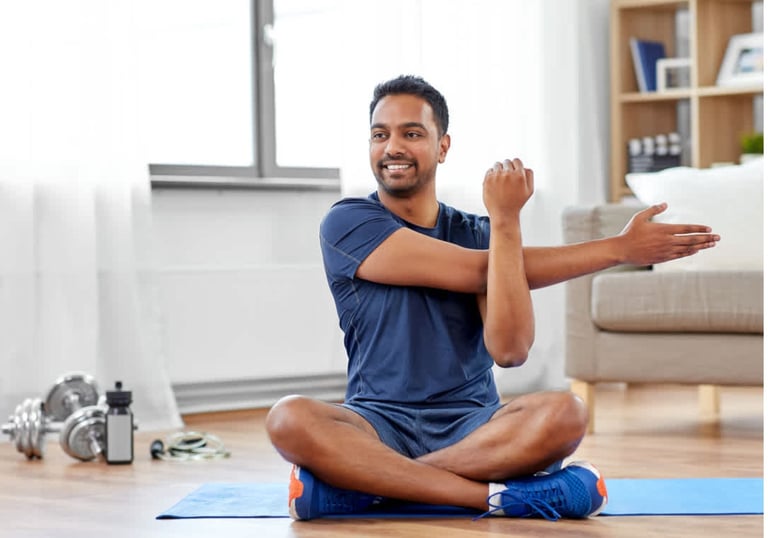 Man sitting cross‑legged while stretching his right arm across his chest, using his left arm to gently support the stretch.